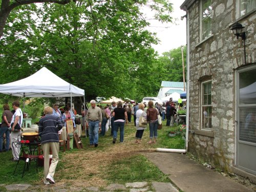 A bustling outdoor market with people browsing stalls under white tents, surrounded by greenery and a stone building.