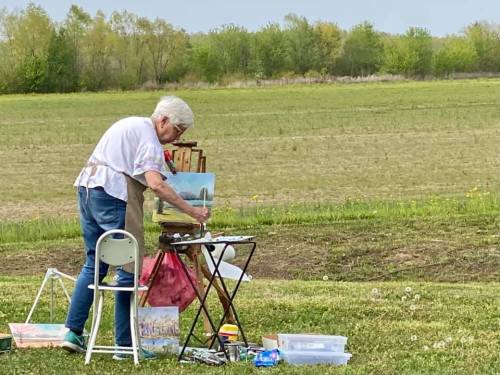 An elderly woman paints on an easel outdoors, surrounded by a grassy field and trees in the background.