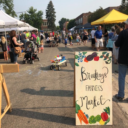 A bustling farmers market with tents, vendors, and shoppers, featuring a colorful sign for "Brookings Farmer's Market."