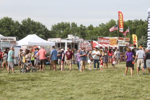 A lively outdoor festival scene with food stalls and a crowd enjoying the event on a sunny day.