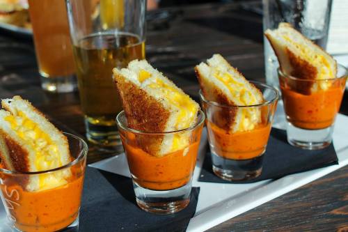 Four grilled cheese sandwiches served in shot glasses with a side of tomato soup, accompanied by a glass of beer.