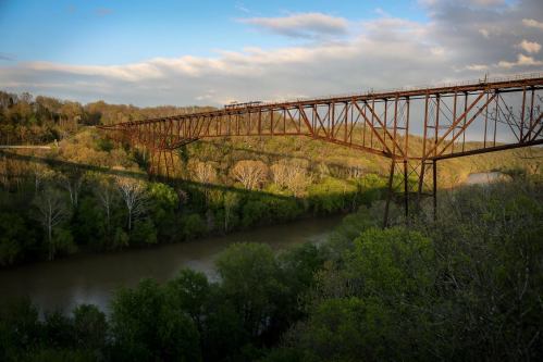A rusted bridge spans a river, surrounded by lush greenery and trees under a partly cloudy sky.
