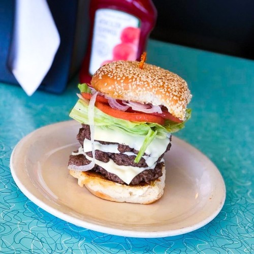 A tall, layered burger with lettuce, tomato, onions, and multiple beef patties, served on a sesame seed bun.