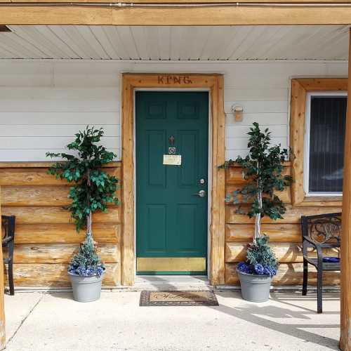 A green door with a sign reading "KING," flanked by potted plants, set against a wooden wall and a concrete path.