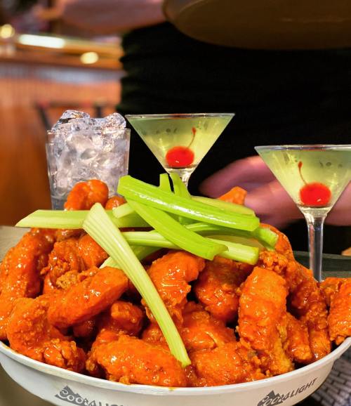 A bowl of spicy chicken wings topped with celery, alongside two cocktails with cherries and a glass of ice.