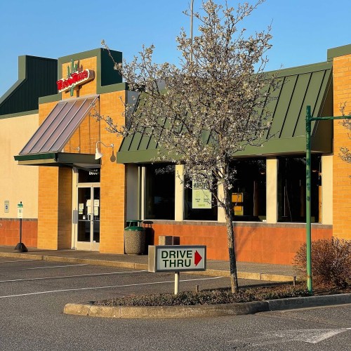 A fast-food restaurant with a drive-thru sign and a tree in front, set in a parking lot on a sunny day.