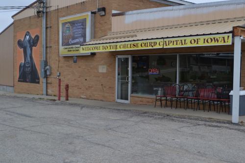 A storefront with a sign reading "Welcome to the Cheese Curd Capital of Iowa!" and a mural of a cow on the side.