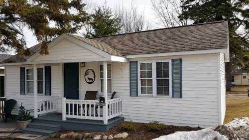 A small white house with a front porch, blue shutters, and a circular wall decoration, surrounded by grass and trees.