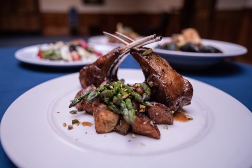 A plate featuring lamb chops with asparagus and roasted potatoes, set against a blurred background of salads.