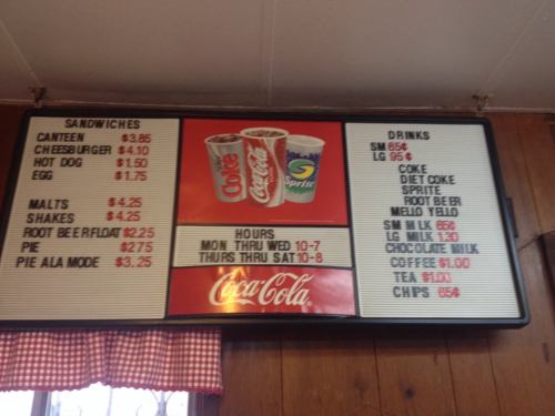 Menu board displaying food and drink prices, including sandwiches, malts, shakes, and various beverages.