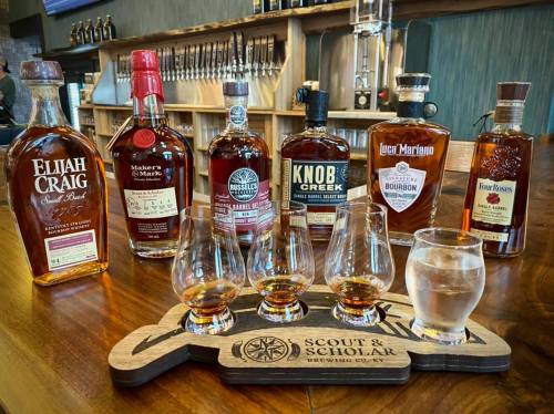 A selection of whiskey bottles with tasting glasses on a wooden tray at a bar.