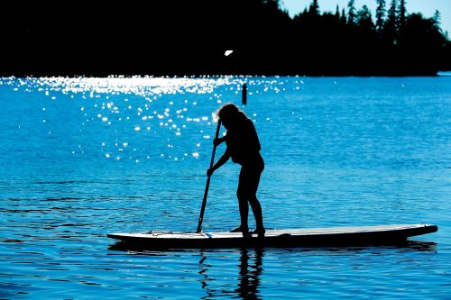 A silhouette of a person paddleboarding on a sparkling lake with trees in the background.