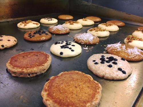 A variety of pancakes cooking on a griddle, topped with nuts, coconut, and berries.