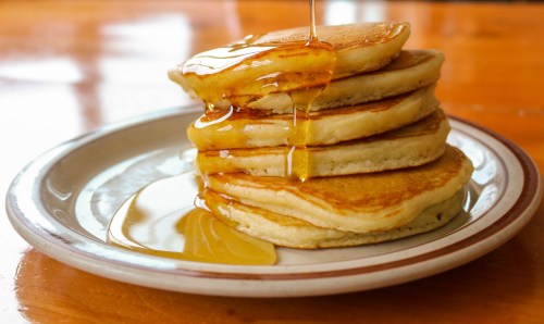 A stack of fluffy pancakes drizzled with golden syrup on a white plate, resting on a wooden table.