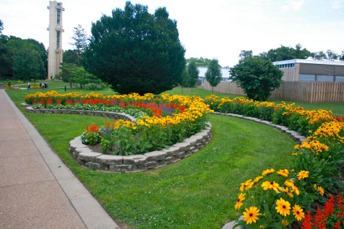 A vibrant flower garden with yellow and red blooms arranged in a spiral pattern, surrounded by green grass and trees.