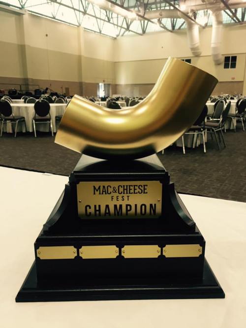 A golden trophy shaped like a macaroni noodle, labeled "Mac & Cheese Fest Champion," on a black base.