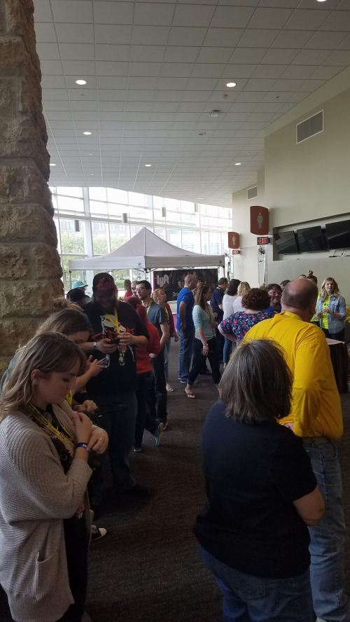 A diverse group of people standing in line at an event, with booths and a large window in the background.