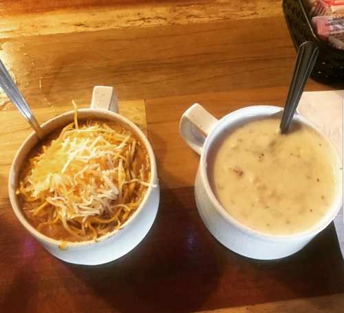 Two bowls on a wooden table: one with cheesy chili and the other with creamy soup, both with spoons.