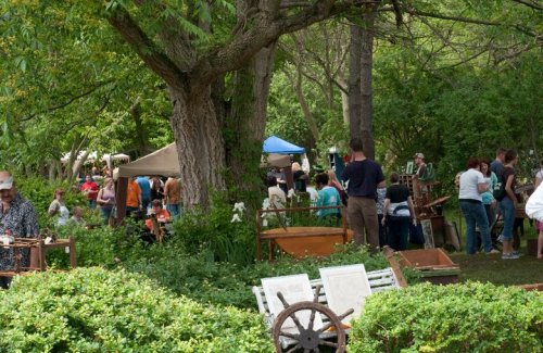 A bustling outdoor market scene with people browsing among trees and tents, surrounded by greenery and wooden displays.
