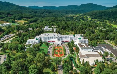 Aerial view of a large hotel surrounded by lush greenery and mountains, featuring a landscaped garden in front.