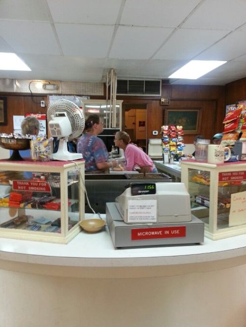 A small diner counter with snacks and a microwave, featuring two people interacting behind the counter.