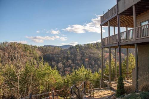 A scenic view from a balcony overlooking a forested valley and distant mountains under a clear blue sky.