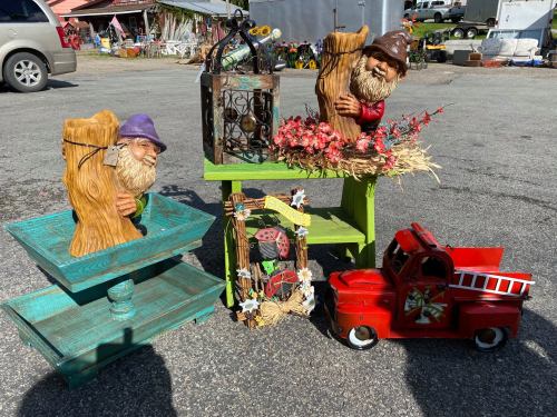 Two whimsical garden gnomes on display with colorful wooden shelves, a vintage truck, and decorative items at a market.