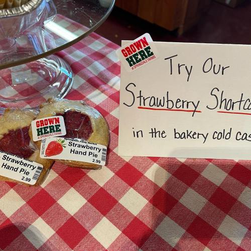 A display of strawberry hand pies with price tags on a checkered tablecloth, alongside a sign promoting strawberry shortcake.
