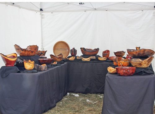 A display of various handcrafted wooden bowls and art pieces arranged on black tables under a white tent.