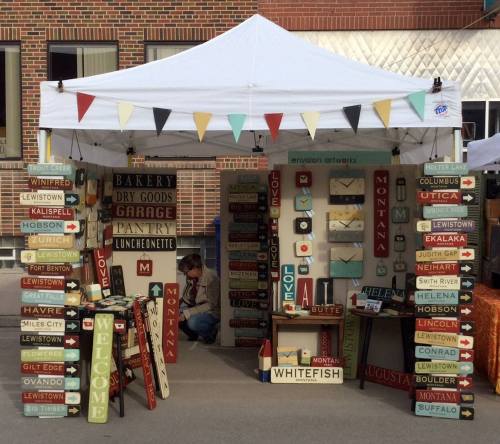 A vendor booth with colorful signs displaying various locations and messages, under a white tent with decorative bunting.