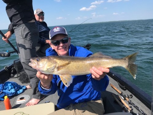 A man in a blue jacket holds a large fish while fishing on a boat, with another person in the background.