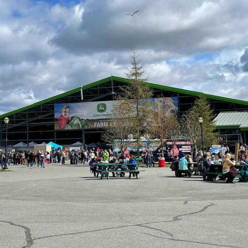 A bustling outdoor market scene with people gathered near a large building featuring a John Deere banner.