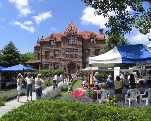 A lively outdoor market with people browsing stalls in front of a large historic building under a blue sky.