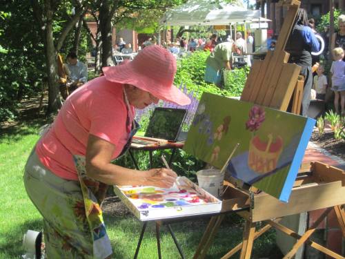 An artist in a pink hat paints on an easel outdoors, surrounded by greenery and a crowd at a fair.