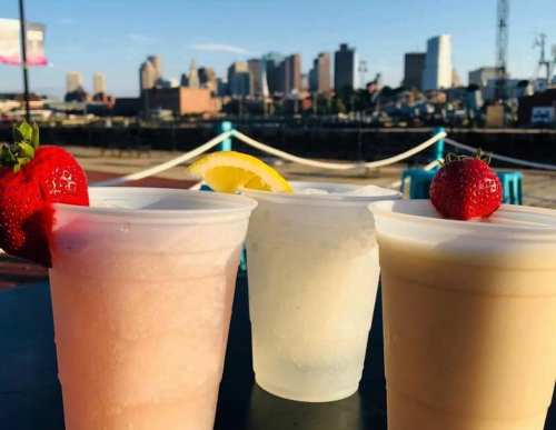 Three colorful drinks in plastic cups with strawberries and lemon, set against a city skyline in the background.