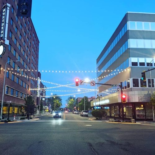 A city street at dusk, featuring buildings, traffic lights, and string lights overhead.