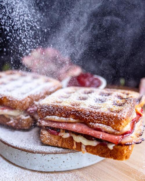 A close-up of a stacked sandwich dusted with powdered sugar, with a blurred background of more sandwiches and a bowl of jam.