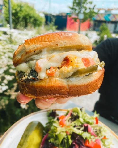 A hand holding a sandwich filled with vegetables and sauce, with a side of salad and pickles in the background.
