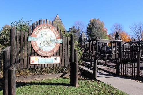 Sign for Hideaway Park with playground structures in the background, surrounded by trees and a clear blue sky.