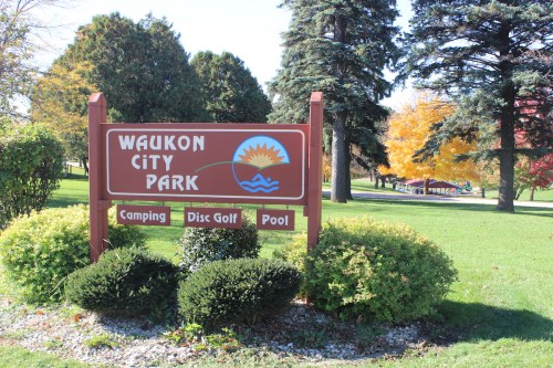 Sign for Waukon City Park, featuring icons for camping, disc golf, and a pool, surrounded by greenery and trees.