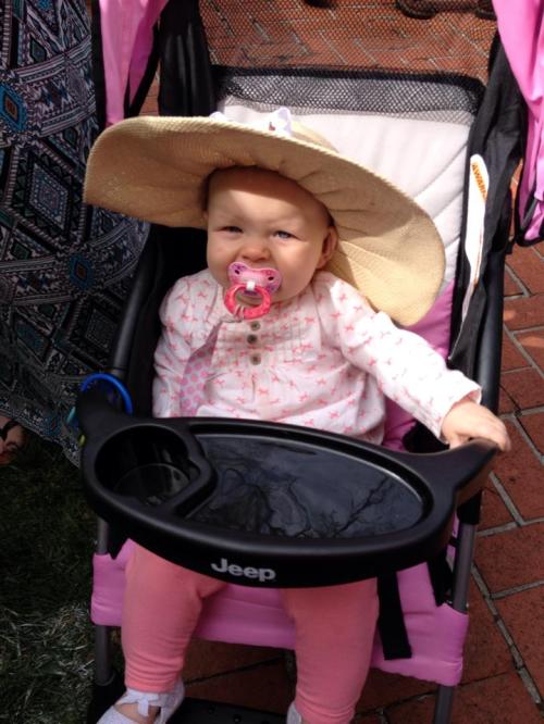 A baby in a pink stroller wearing a large sun hat and a pacifier, sitting outdoors.