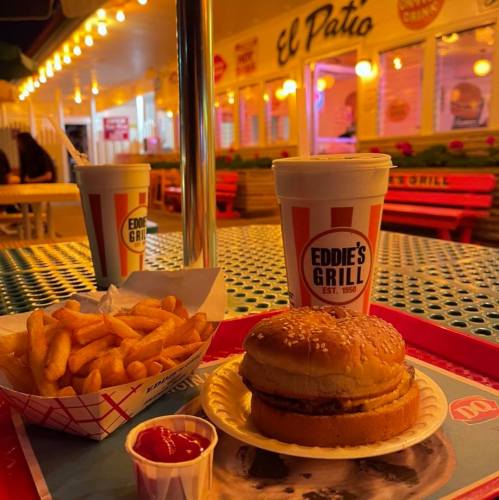 A burger and fries on a table with two drinks, illuminated by warm lights in a casual outdoor dining setting.