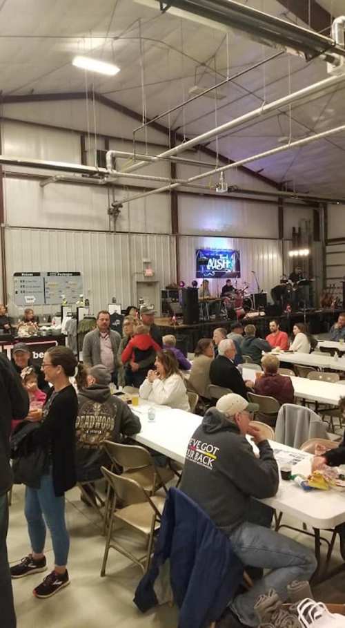 A lively indoor event with people seated at tables, a stage in the background, and a band preparing to perform.
