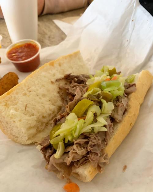 A sandwich filled with sliced roast beef, lettuce, and pickled peppers on a soft hoagie roll, with a side of dipping sauce.