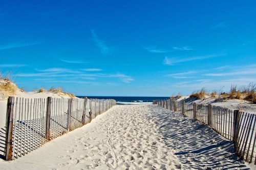 A sandy path leads to the ocean, bordered by wooden fencing and grass under a clear blue sky.