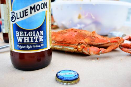 A bottle of Blue Moon Belgian White beer next to a cooked crab and a bottle cap on a table.