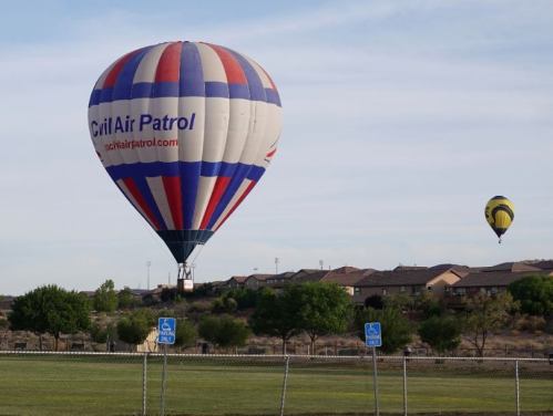 Two hot air balloons in the sky above a grassy field, with houses in the background and a clear blue sky.
