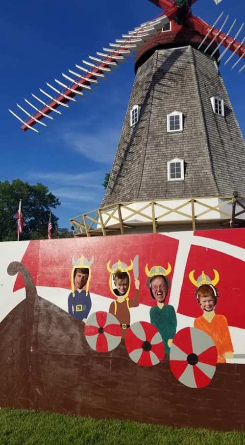Three people with Viking helmets pose in front of a painted Viking ship and a windmill on a sunny day.