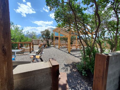 A sunny outdoor space with wooden structures, gravel paths, and trees, surrounded by mountains and blue skies.
