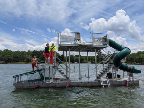 Three people on a floating platform with a slide in a lake, surrounded by trees and a blue sky with clouds.